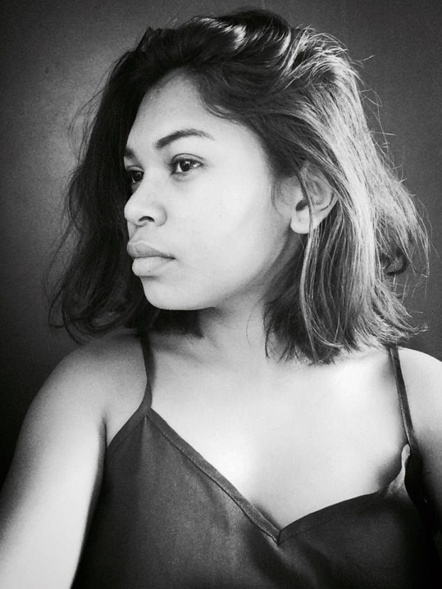 Eine selbstbewusste Frau in einem schwarzen Kleid posiert in einem Schwarz-Weiß-Foto, ihr glatter Bob und das dezente Make-up unterstreichen ihre selbstbewusste Haltung an der Wand.