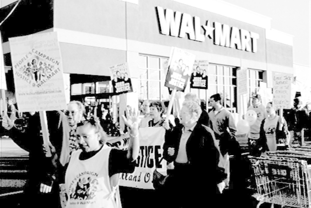 Schwarz-weißes Bild von Menschen, die draußen protestieren und Schilder halten, mit einem Einkaufszentrum im Hintergrund.