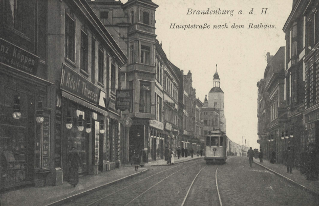 Schwarz-weiß-Foto einer Straßenkreuzung in Brandenburg, Deutschland mit einer Tram, Passanten, Gebäuden, Schildern, Straßenlaternen und einem bewölkten Himmel.