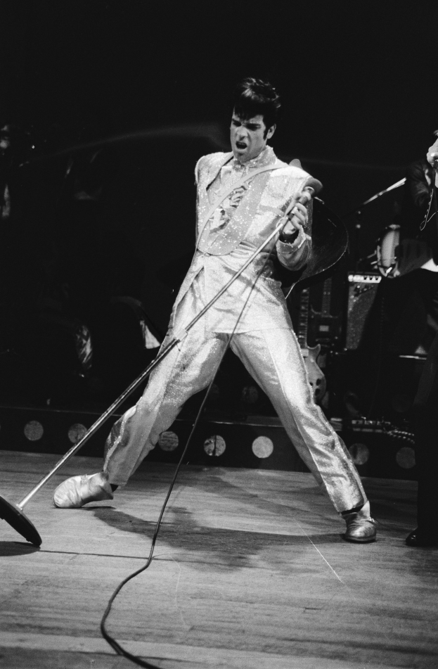 Elvis Presley in einem weißen Jumpsuit auf der Bühne des Apollo Theaters in Las Vegas, mit einem anderen Musiker rechts von ihm und Bühneninstrumenten und Lichtern im Hintergrund.